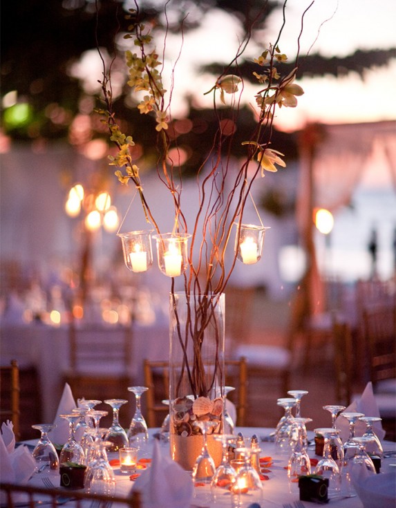Tea light candles hanging from a twig centerpiece on a white linen table at dusk.