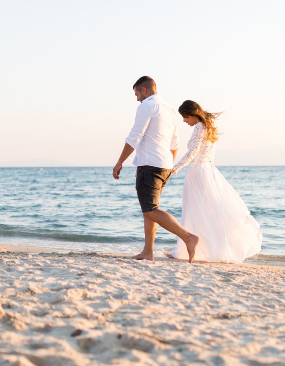 Woman wearing white dress holding hands with a man while walking down the beach at sunset.