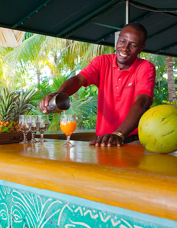 Male bartender wearing a red polo shirt pouring an orange cocktail into a stemmed glass.
