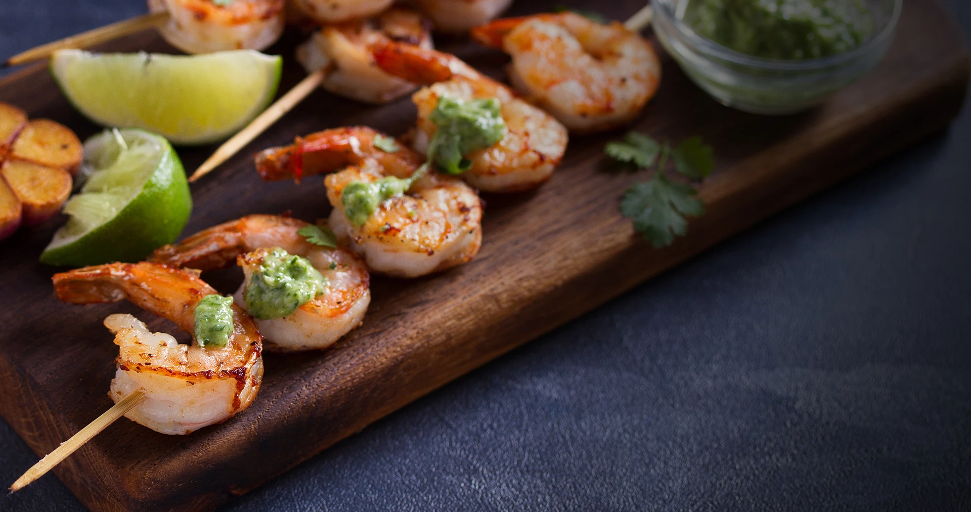 Grilled shrimp skewers served with green finishing sauce on a brown platter.