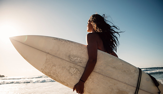 Woman with long brown hair holding a white surfboard walking along the beach at sunset.