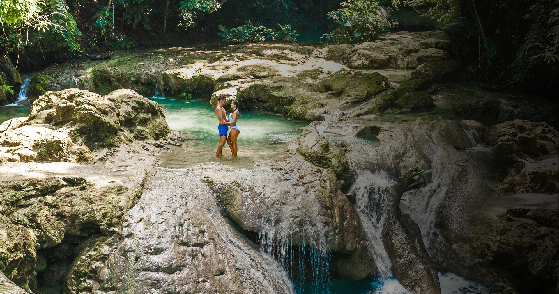 Man and woman hugging while standing on rocks next to a waterfall.