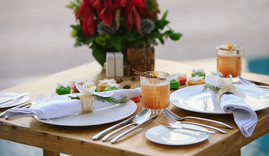 Light oak colored table setup for dinner with two cocktails served in a rocks glass at dusk.