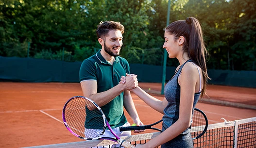 Male and female shaking hands on the dark orange tennis court with the sun shining from above.