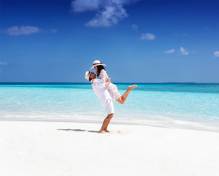 Man and woman wearing white sun hats on the beach next to the clear blue water.