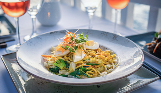 A large bowl of sauteed noodles topped with eggs and scallions served with a glass of rose.