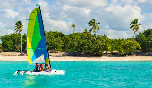 Neon blue, green, and yellow sailboat with three people on it in the blue water.