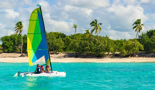 Neon blue, green, and yellow sailboat with three people on it in the blue water.