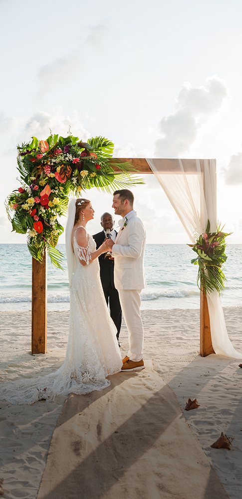 a man and woman in white suit and white suit standing under a floral arch on a beach