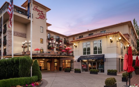 The exterior of Inn at the 5th, a luxury boutique hotel located in Eugene, Oregon’s 5th Street Public Market. The hotel’s elegant façade features balconies adorned with flower boxes and red patio umbrellas, with Pendleton Woolen Mills retail store on the 