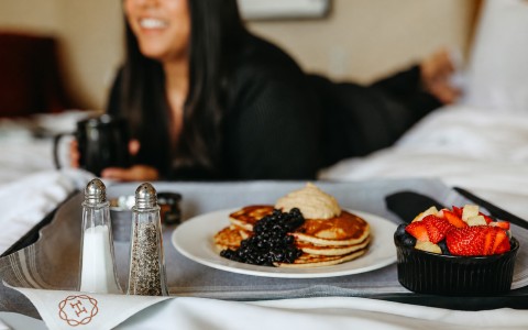 A woman relaxing with a cup of coffee on a bed and Hemlock room service pancakes