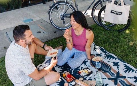 A man and woman sitting on a Pendleton picnic blanket enjoying charcuterie food and drinks on a patch of grass that they biked to