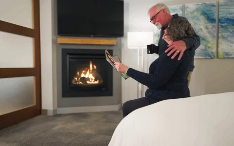 a man and woman sitting on a bed in front of a fireplace looking at a magazine
