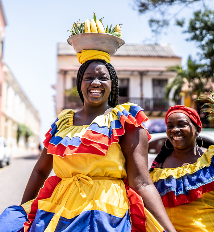 a group of women wearing colorful dresses and a bowl of bananas on their head