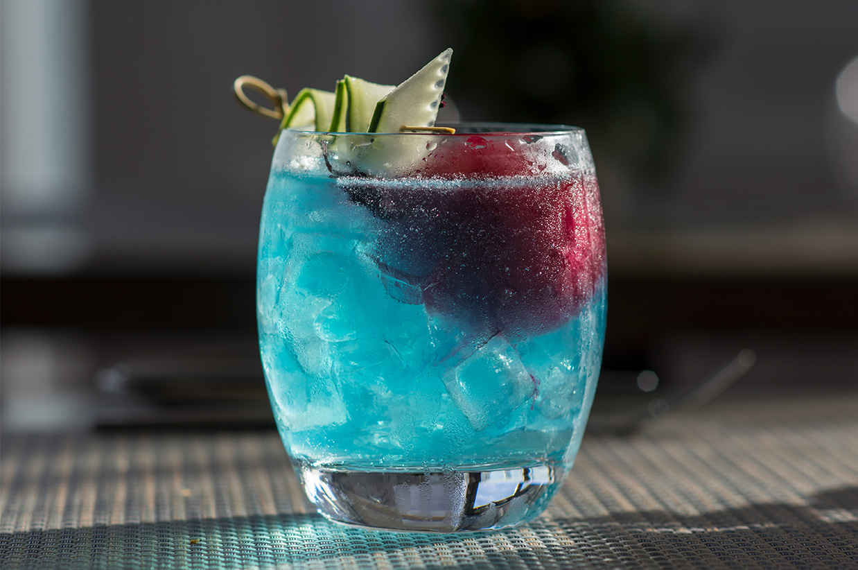 a blue drink with cucumber and ice in a glass