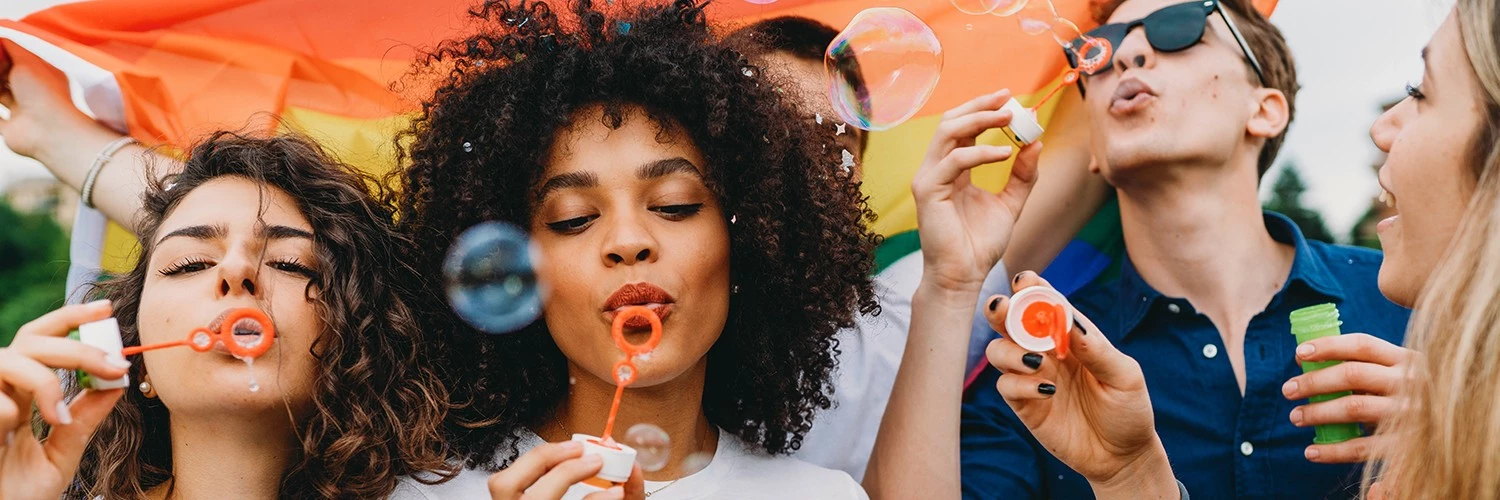 a group of friends blowing bubbles and laughing with a pride flag behind them