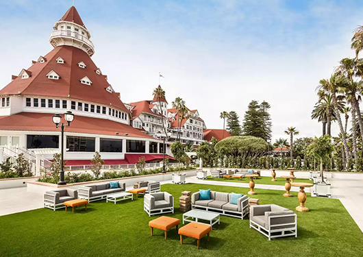 a large building with a large round structure and a lawn with chairs and tables with Hotel del Coronado in the background