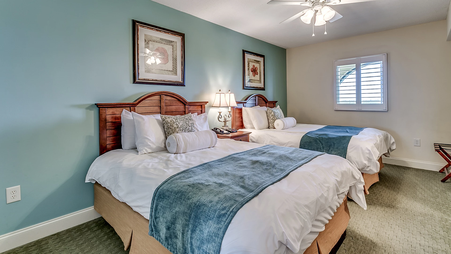 A hotel room with two twin beds, blue accent wall, wooden headboards, and a ceiling fan.