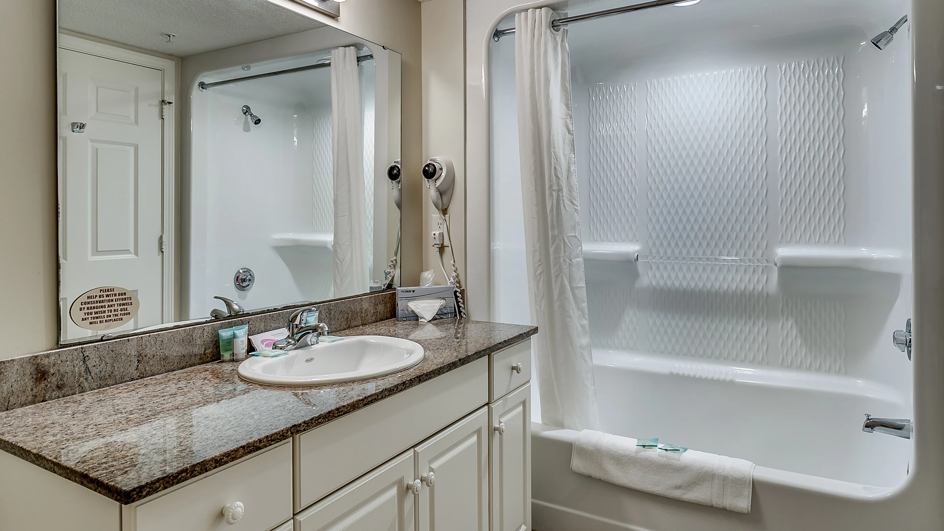 A clean bathroom with a granite countertop, white cabinets, and a large mirror over the sink, with a tub/shower combo to the right.