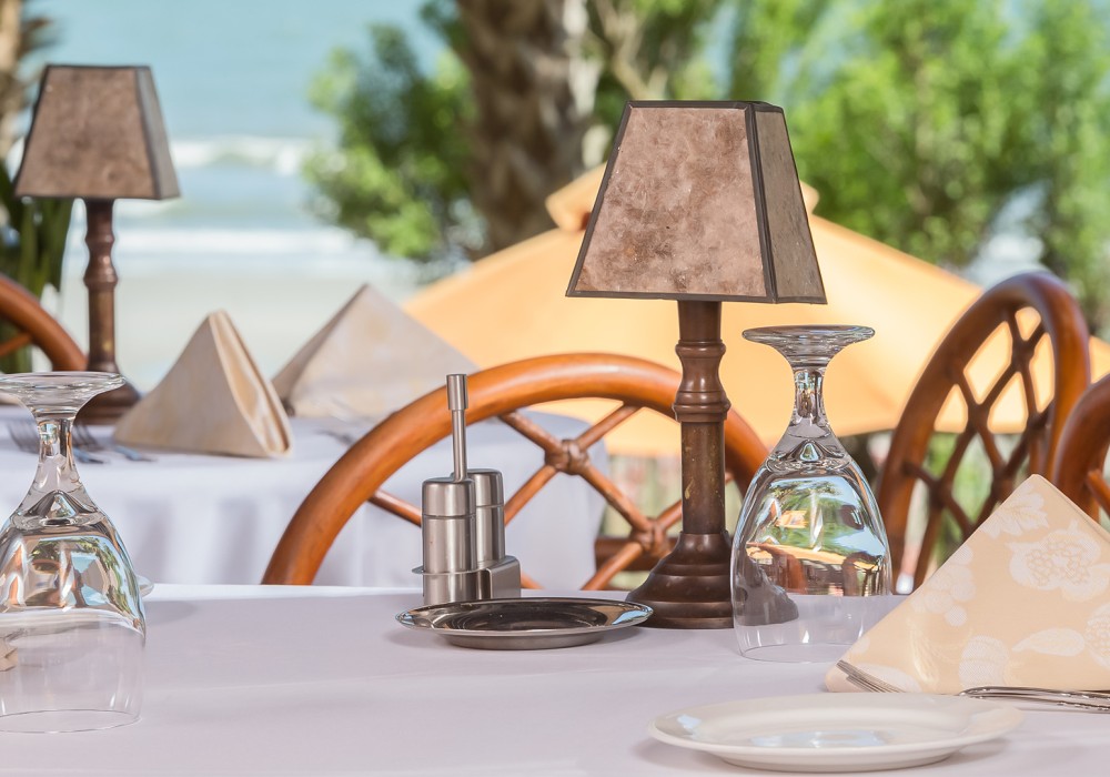 A dining table set with white tablecloth, glasses, napkins, and lamps, with a blurred ocean view in the background.