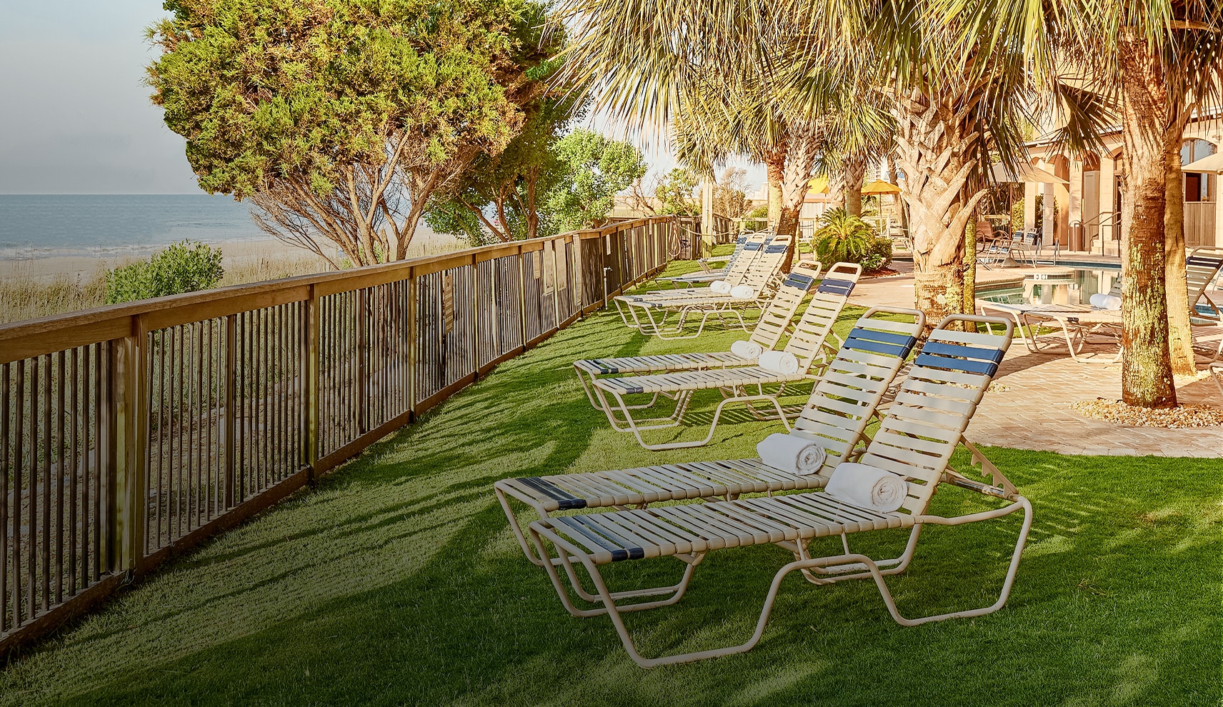 A poolside area with lounge chairs, trees, and a fence overlooking the ocean.
