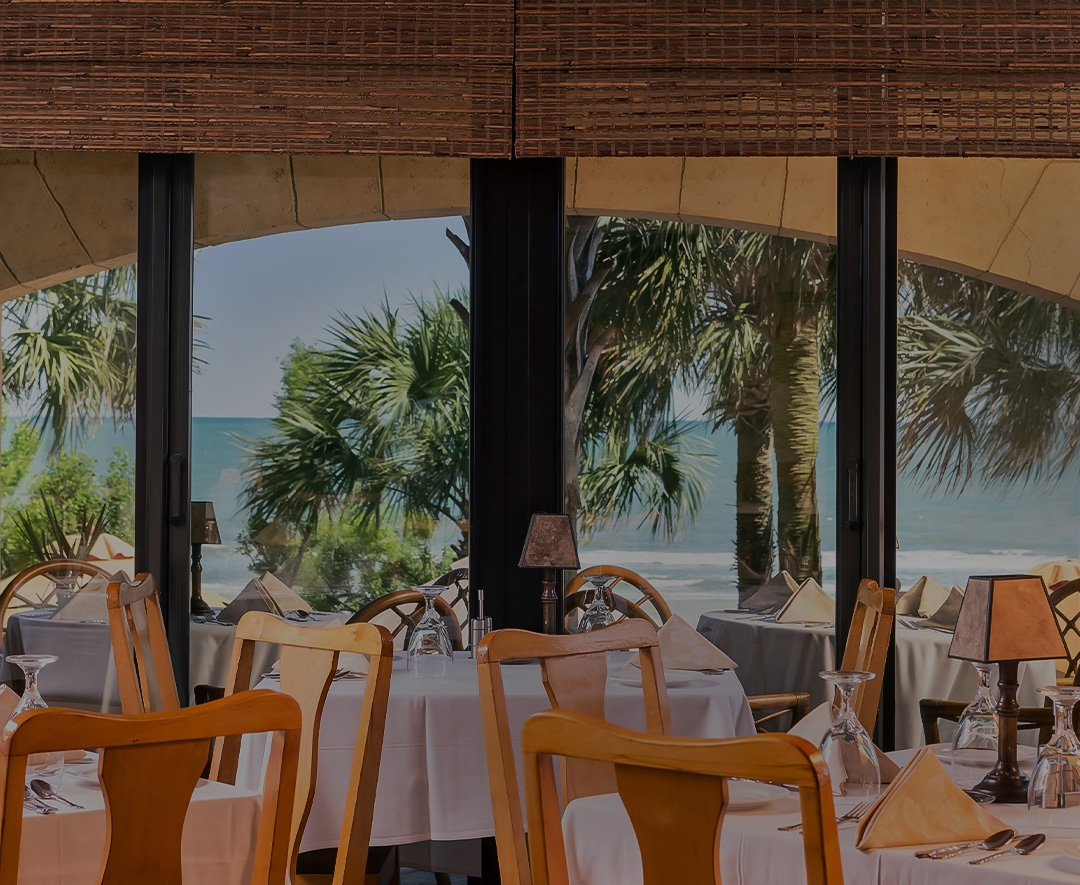 A dining area with white tablecloths and wooden chairs, overlooking the ocean through large windows with palm trees in the background.