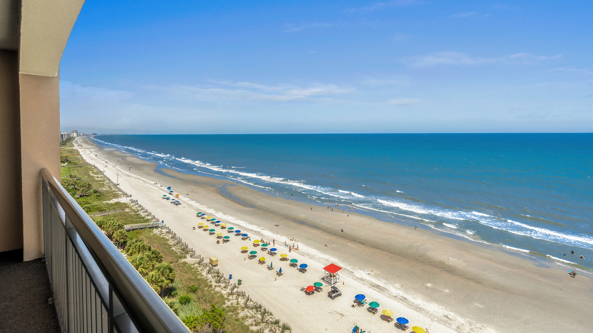 A beach view from a balcony with a railing, overlooking a sandy beach with colorful umbrellas and a vast ocean under a blue sky.