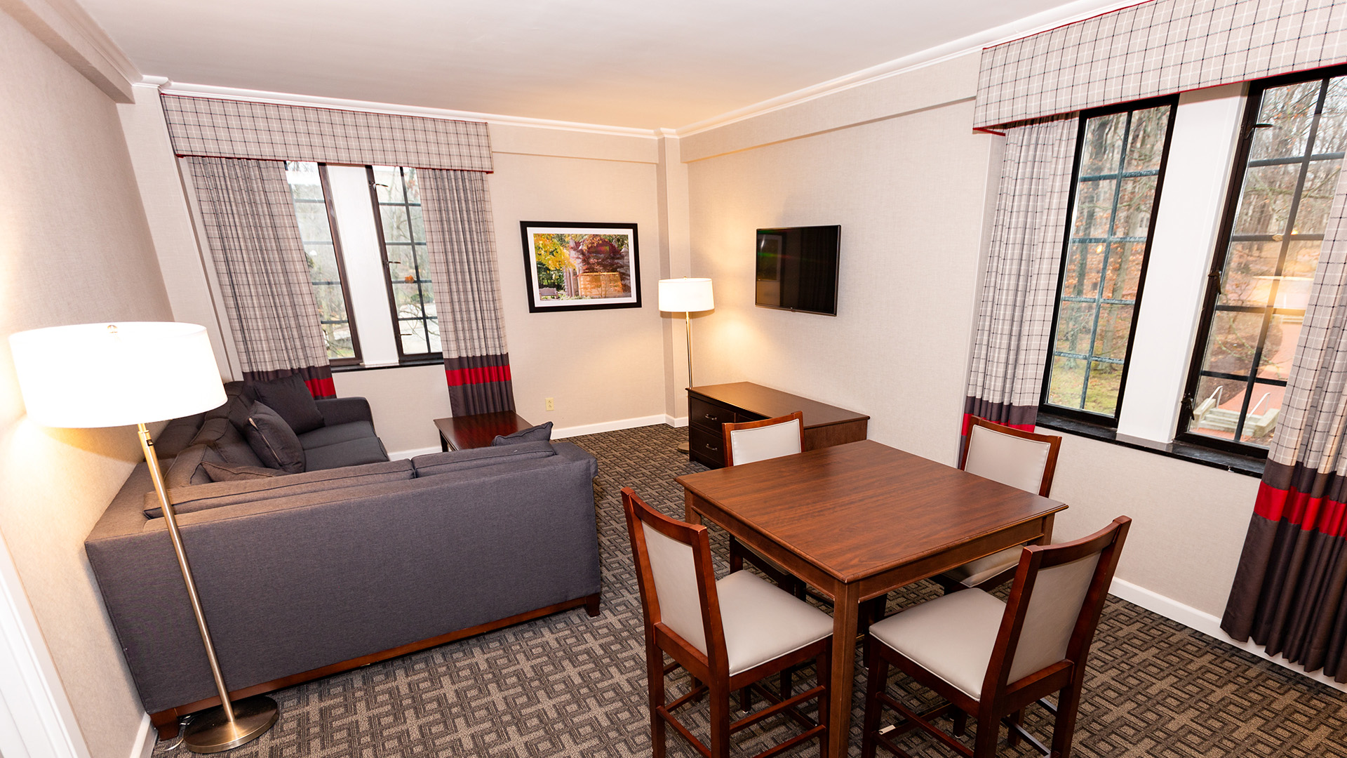 living area with a grey sectional, wall-mounted TV, and a four-person dining table by large windows