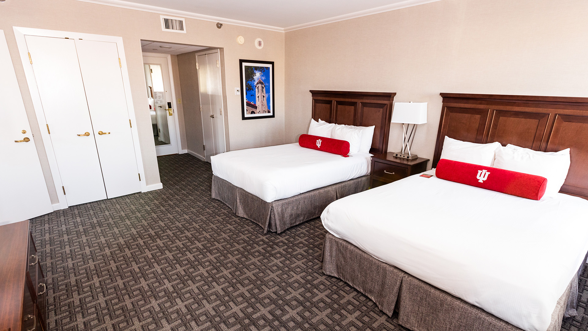 a hotel room featuring two queen beds with red Indiana University pillows and a dark wood headboard