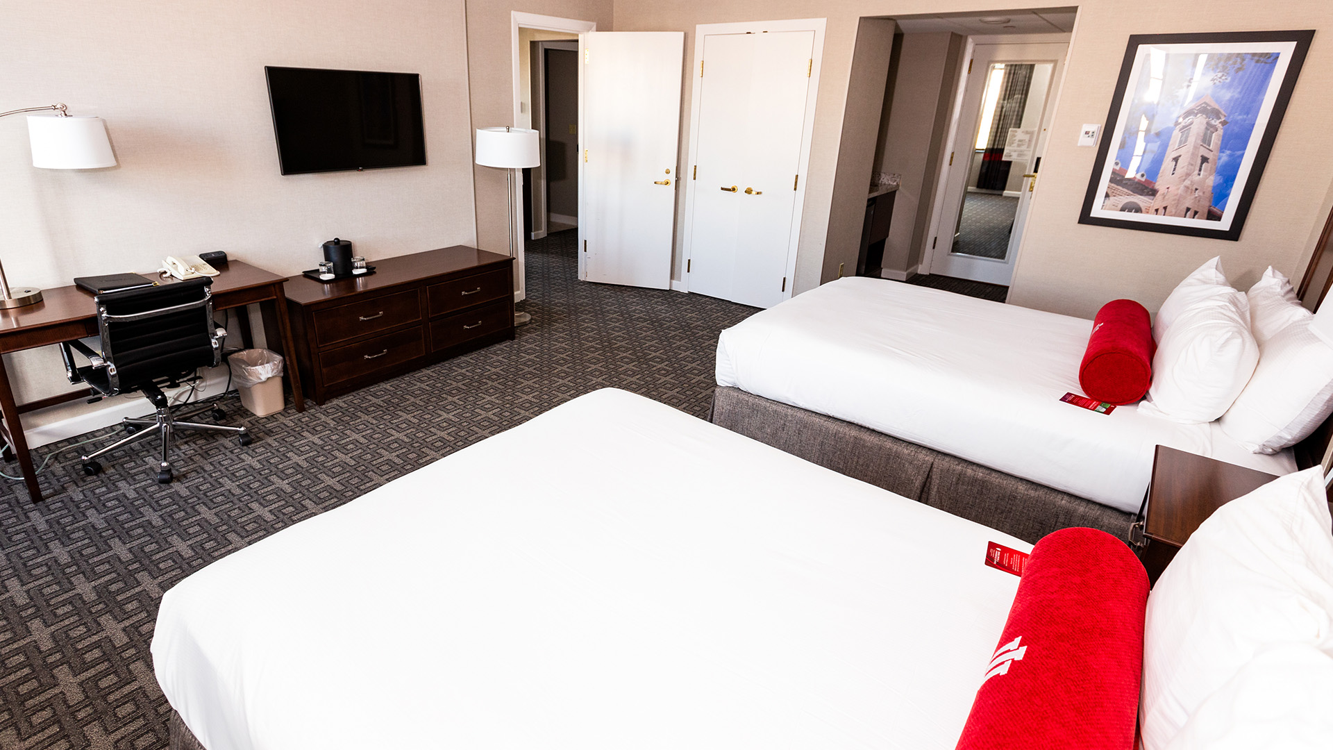 hotel room with two double beds, red bolster pillows, a work desk, TV, and entry to an adjoining space