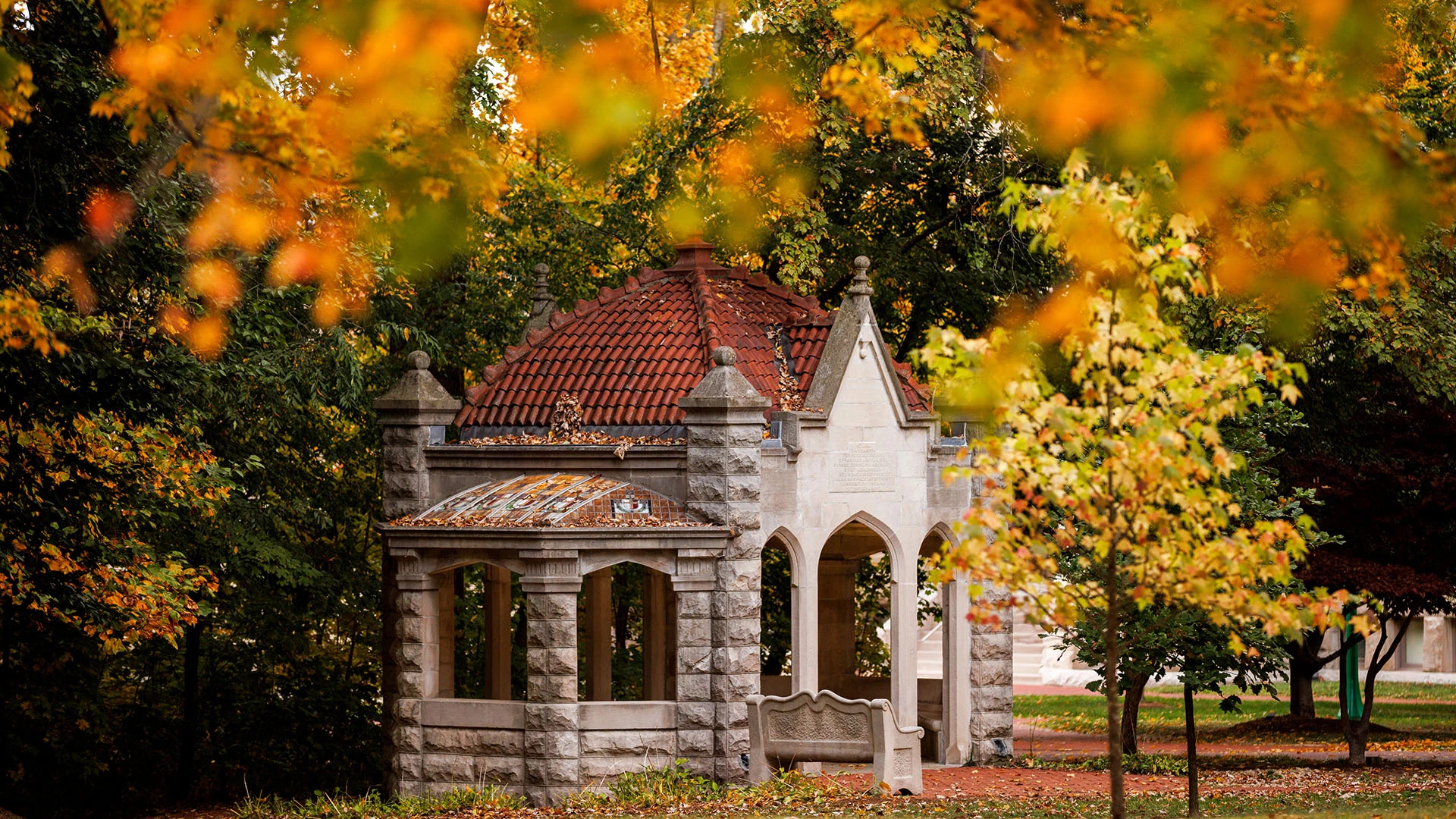 a stone structure with a red roof and a bench in the middle of trees