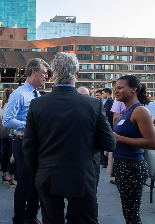 A group of men and women smiling and talking on a rooftop terrace at dusk.