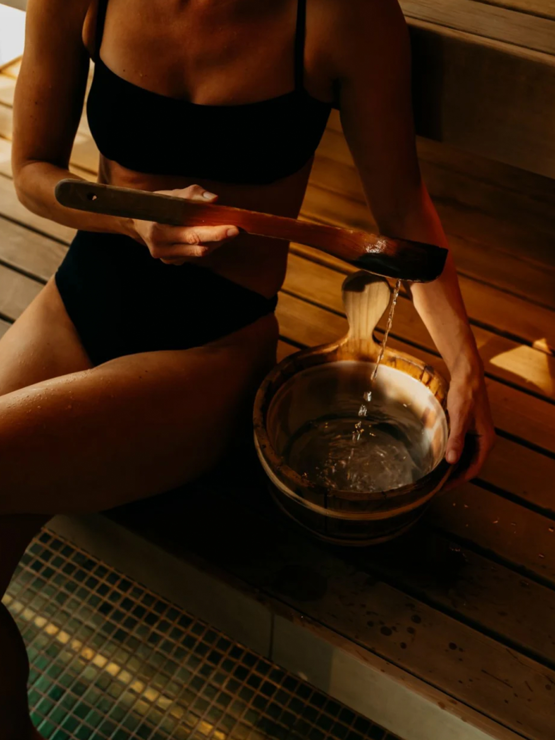 a woman sitting on a bench in a sauna pouring water into a bowl