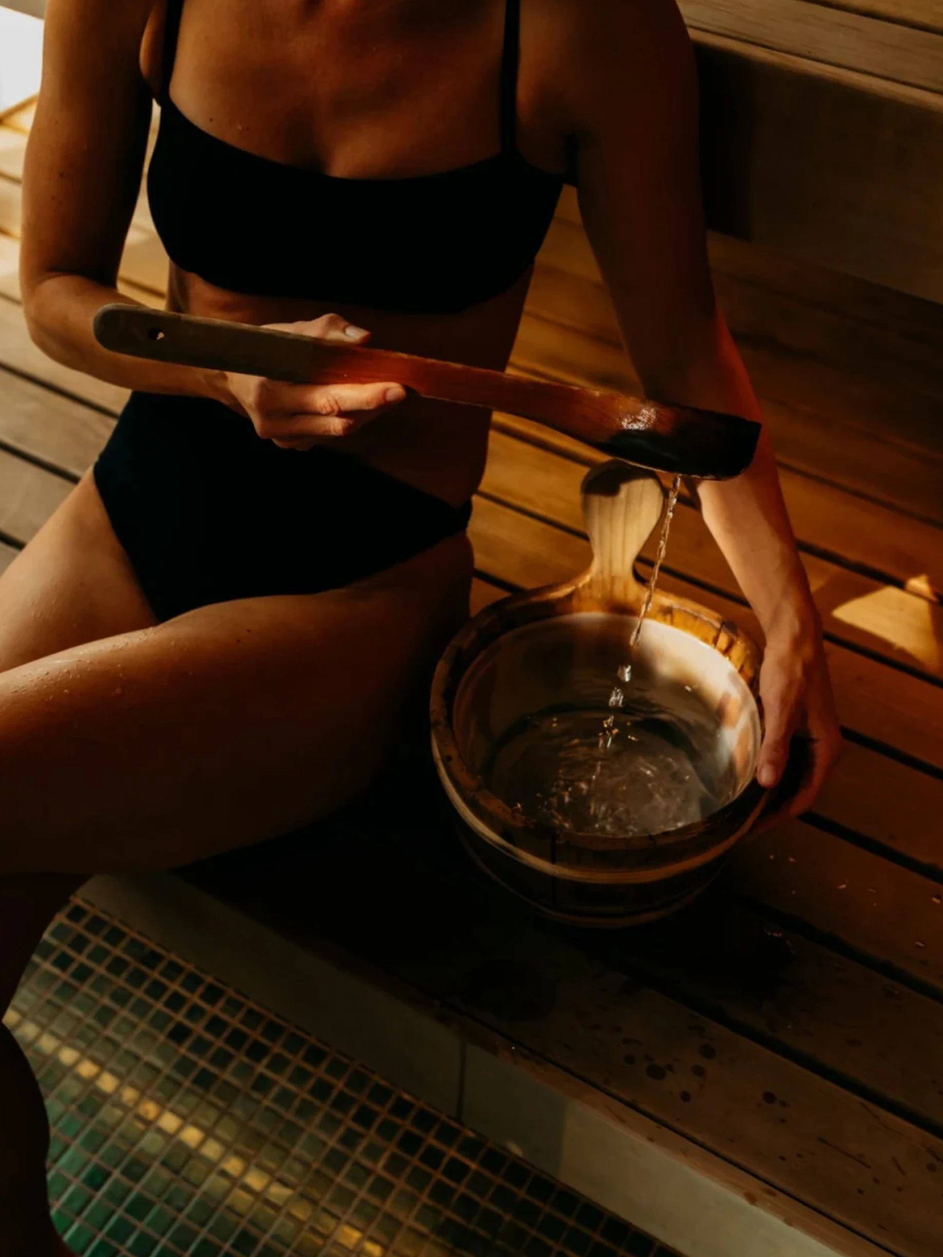 a woman sitting on a bench in a sauna pouring water into a bowl