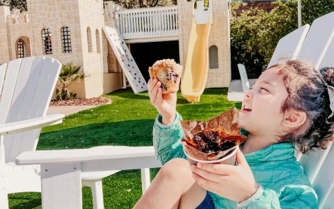 little girl sitting on a white lounge chair enjoying a muffin with a castle playground in the background 