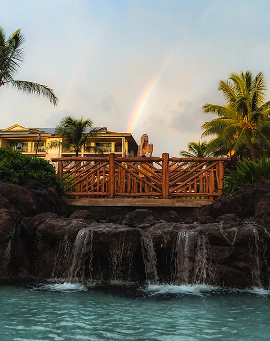 a woman sitting on a bridge over a pool with a rainbow in the background