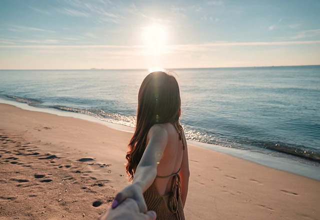 adventurous female leading a male by hand down the sandy beach towards the shore while the sun sets