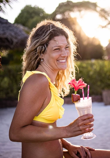 A woman with blonde hair and in a yellow two-piece bikini enjoying a drink by the beach.