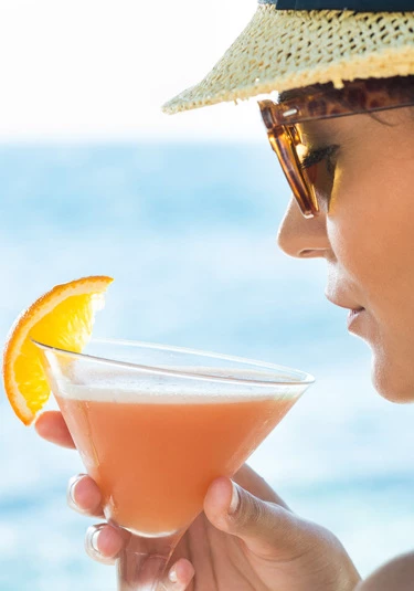 A close up image of a woman with sunglasses and a sunhat enjoying a orange colored drink with a slice of lemon.