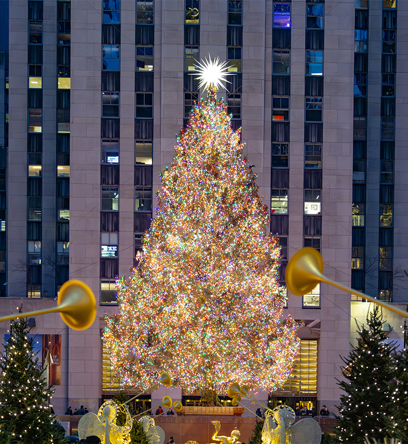 a large christmas tree with lights and a star in the middle with Rockefeller Center in the background