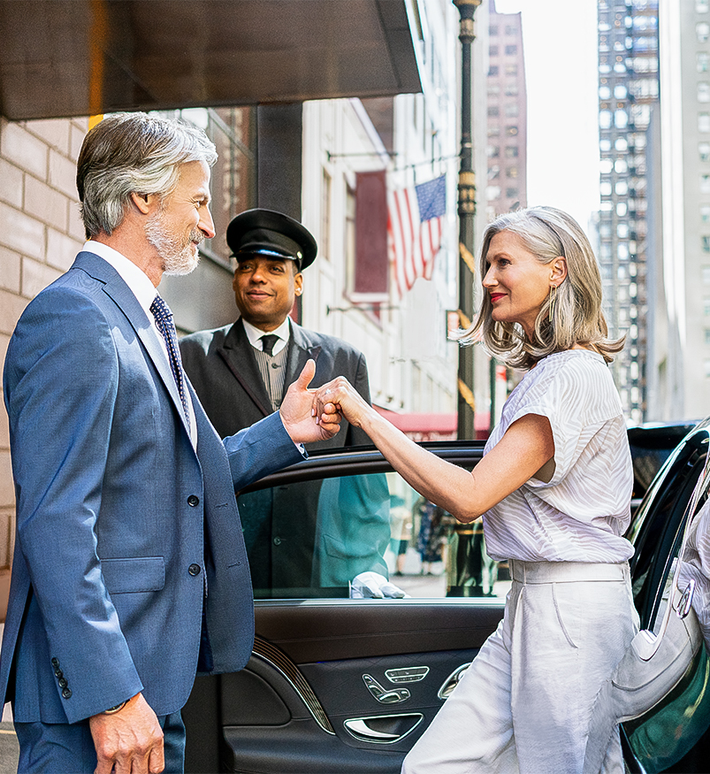 a man in blue suit helping woman in beige linen pant set out of a car with hotel concierge member smiling in the background