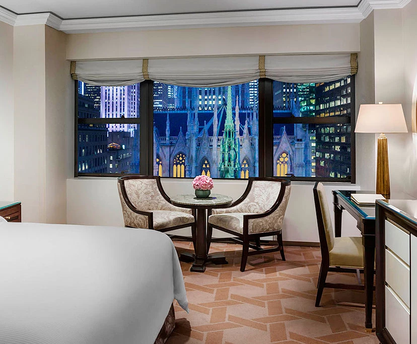 A hotel room with a bed, chairs, and a table in front of a large window showing a city view at night.