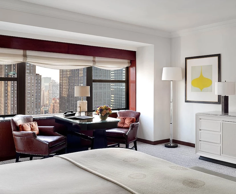 A luxurious hotel room with a city view, featuring a large bed, a desk with two chairs, and a dresser.