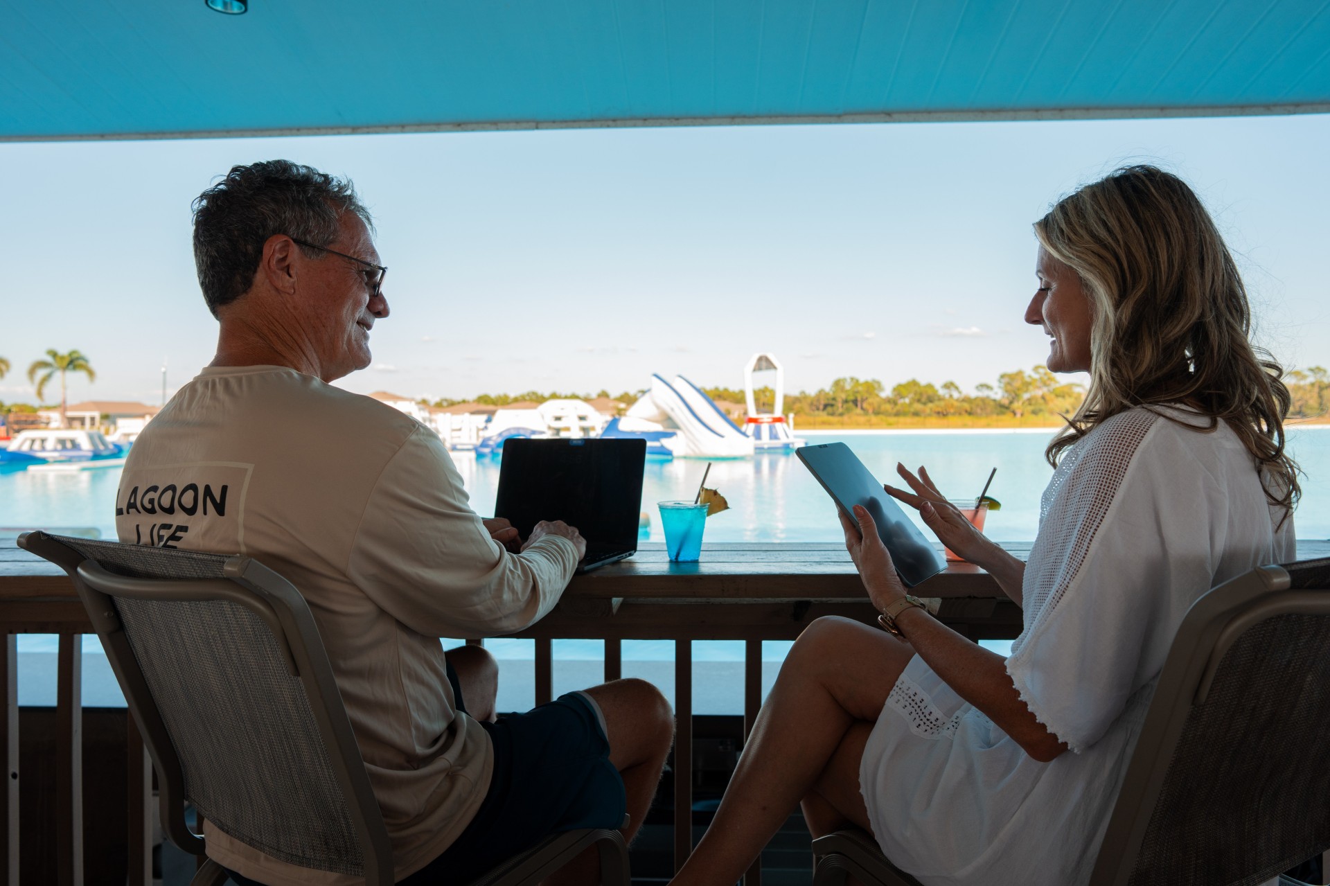 a man and woman sitting at a table with laptops and a boat in the background