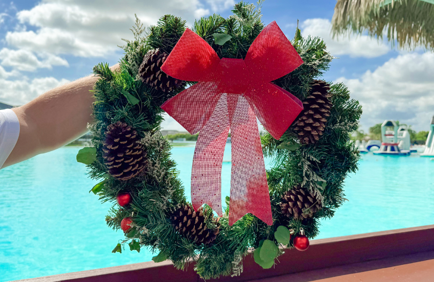 a hand holding a wreath with a red bow and pine cones