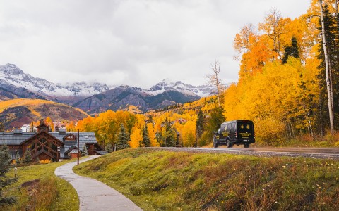 a black van on a road with yellow trees and mountains in the background