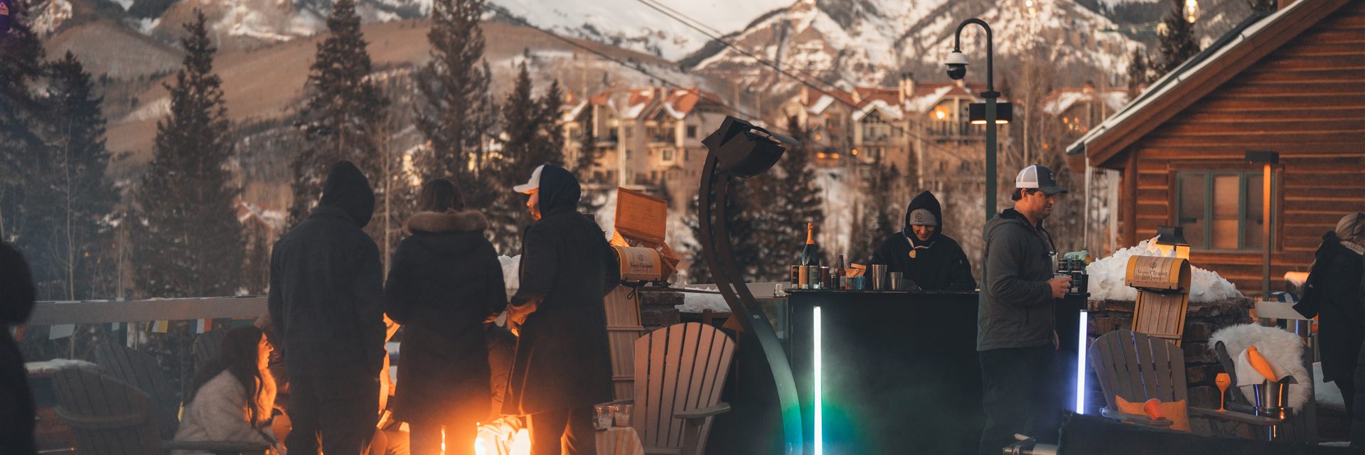 Guests gathered around a firepit outside Mountain Lodge with snowy mountains behind