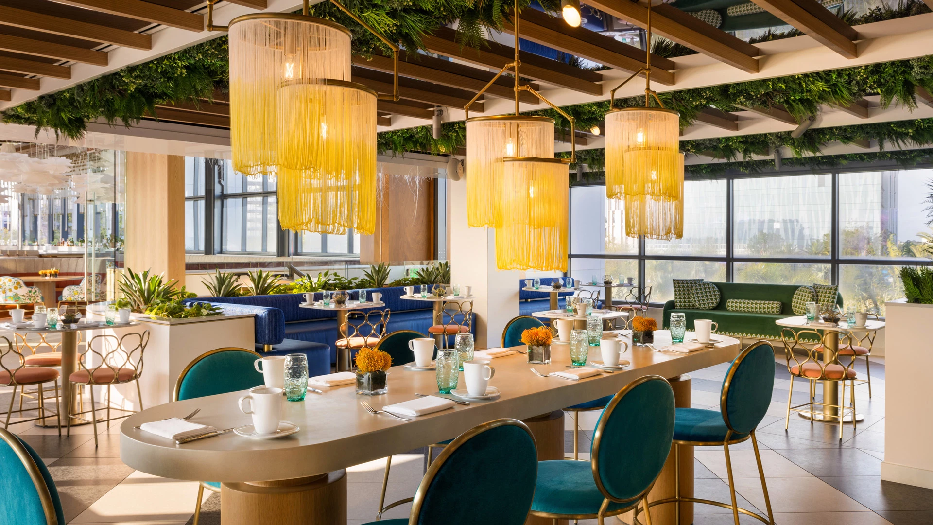 A dining area with a long table set for breakfast, teal chairs, and yellow hanging lights.