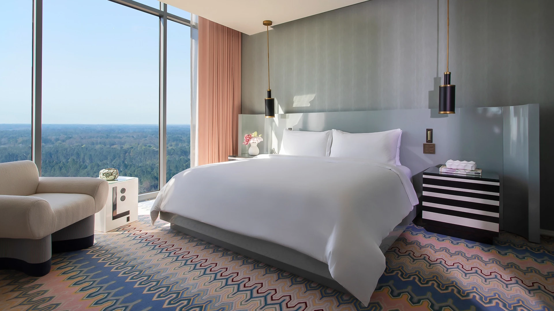 A hotel room with a large window overlooking a city, a king-sized bed, and a comfortable armchair.