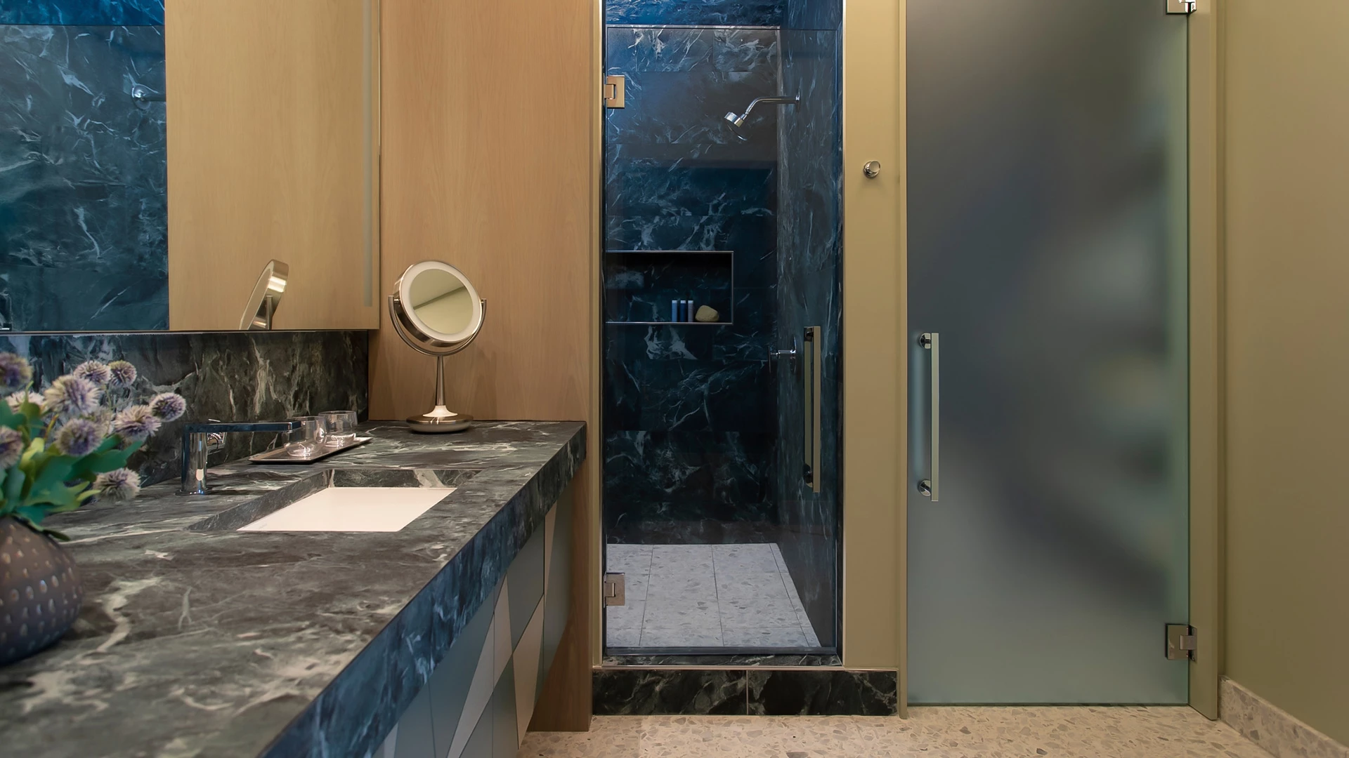 A bathroom with a marble countertop, glass-enclosed shower, and flowers